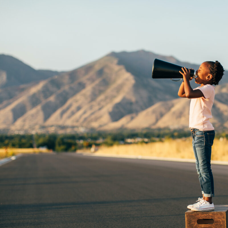 A young retro girl stands on a wooden box while yelling through a megaphone to have her message heard. Image taken in Utah, USA.